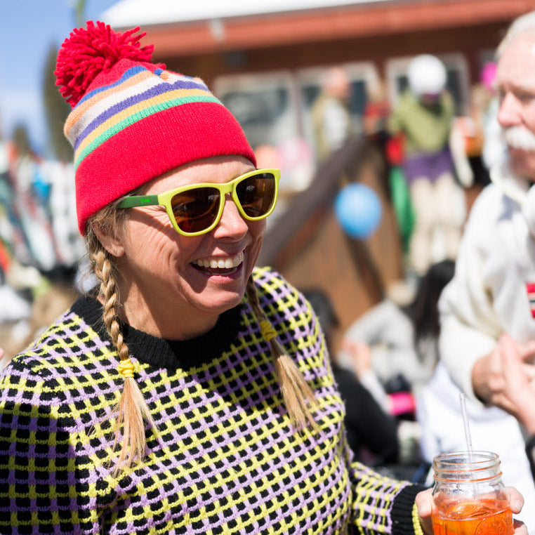 Person wearing a colorful knit hat and sunglasses, holding a drink with a straw, in an outdoor setting.