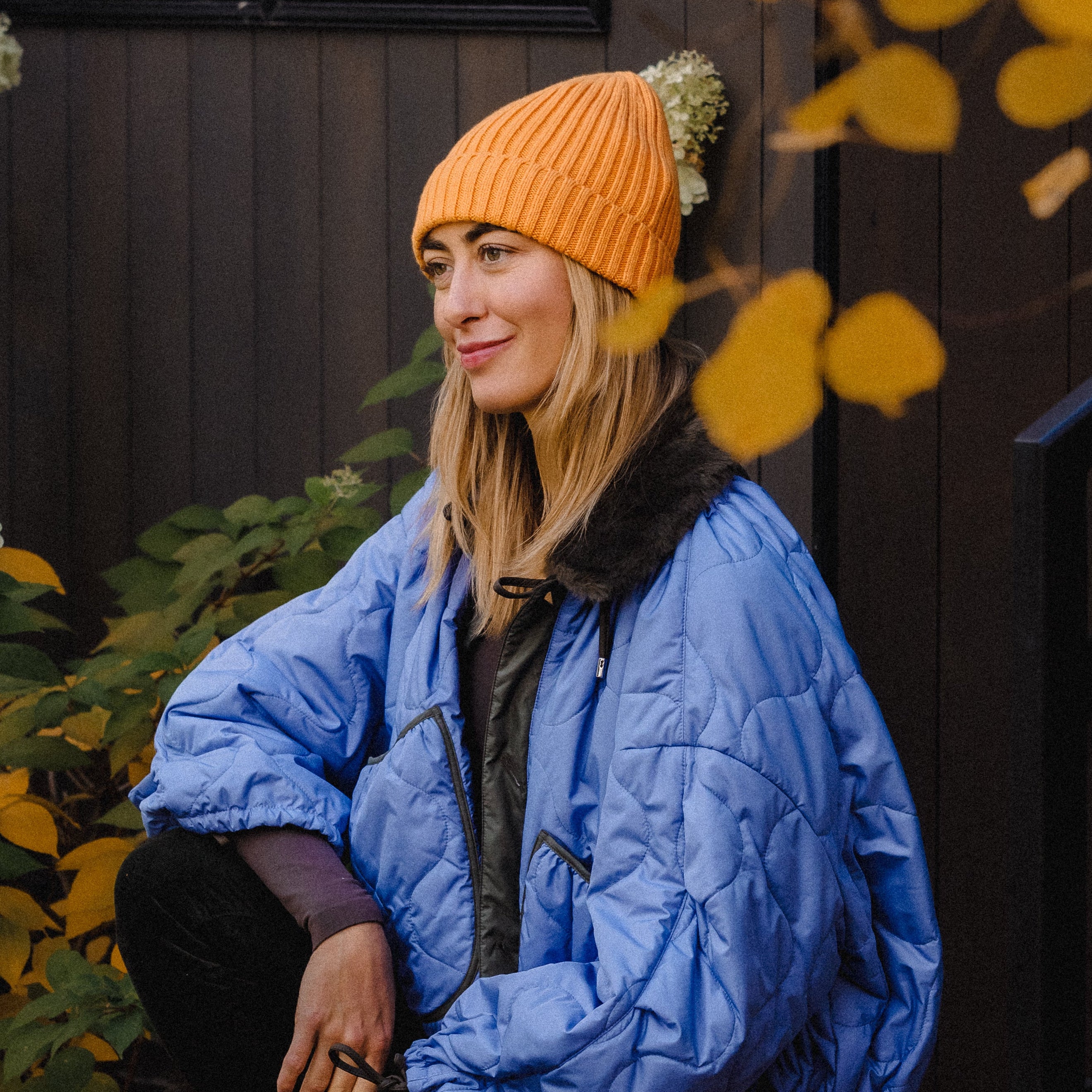 Woman wearing a blue jacket and orange beanie sitting outdoors with autumn leaves and flowers.
