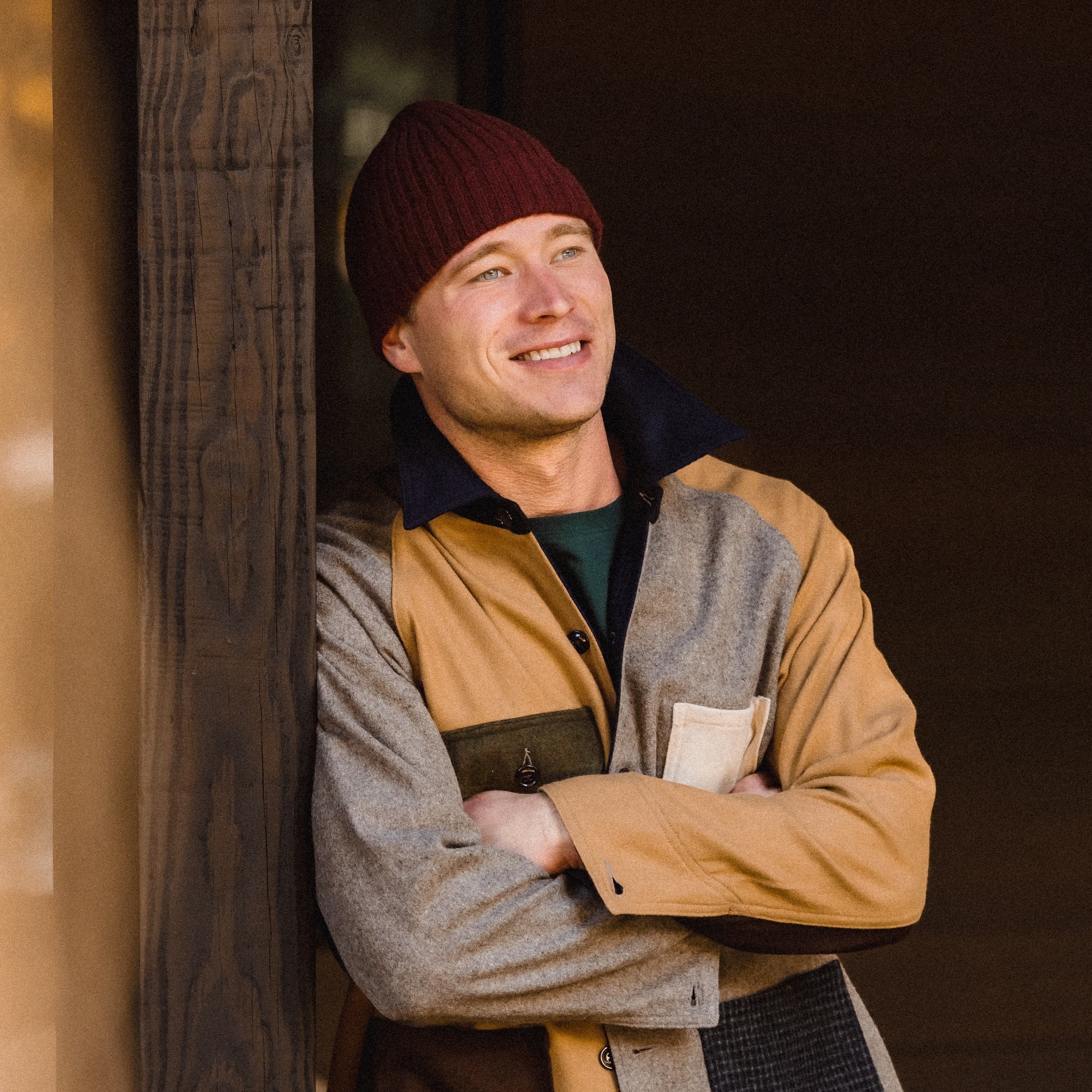 Man wearing a layered outfit with a beanie, jacket, and jeans standing against a wooden wall.