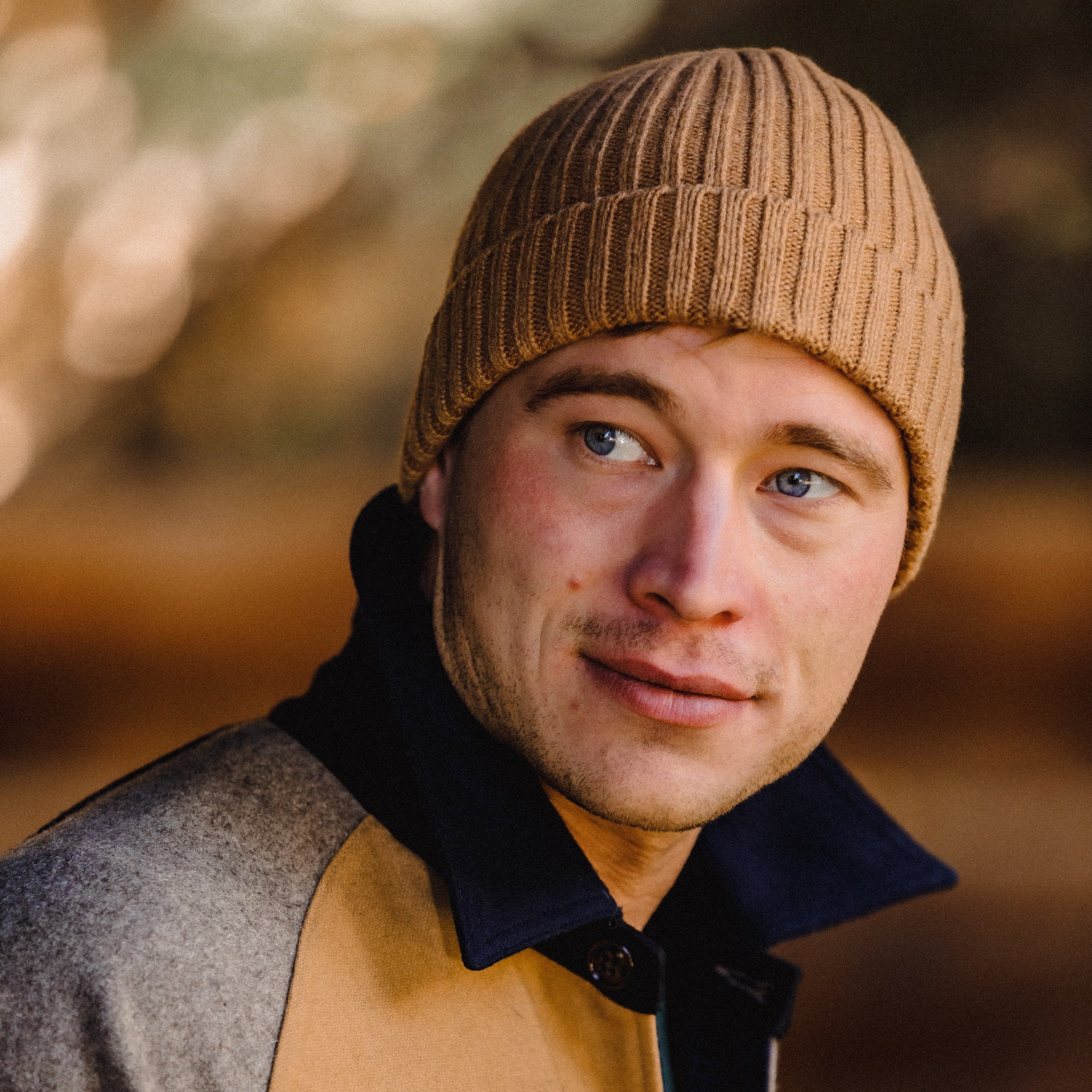 Man wearing a brown knit beanie and a layered outfit with a blurred natural background