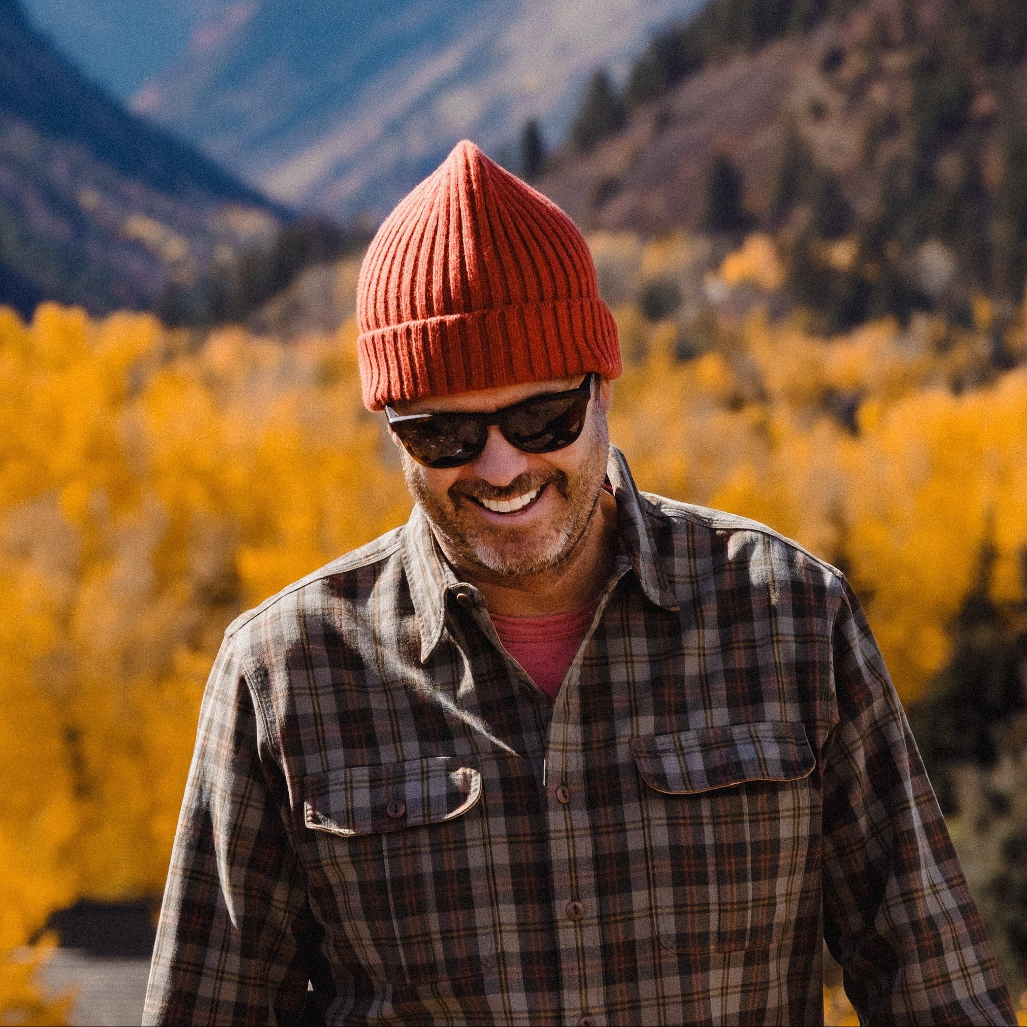 Person wearing a plaid shirt and orange beanie standing in a mountainous area with yellow trees and snow-capped peaks.